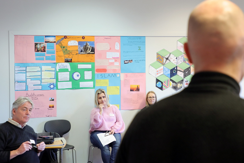 Vi ser tre voksne personer som sitter med ryggen mot en vegg og ser på en mann som vi ser i forgrunnen, med ryggen til.