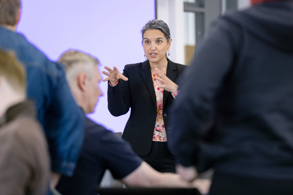 En mørkhåret dame står i midten og gestikulerer mens hun snakker til fire personer som sitter med ryggen til.