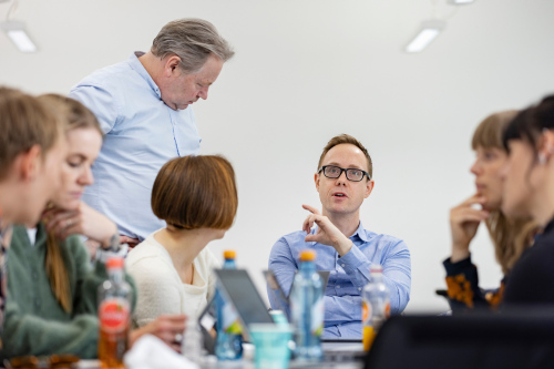 En gruppe som sitter ved et bord og en mann som står til venstre og diskuterer med en mannlig deltager