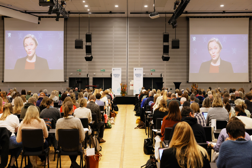 Oversiktsbilde over publikum på Novemberkonferansen. Vi ser Synnøve Mjeldheim Skaar, statssekretær i Kunnskapsdepartementet på to skjermer.