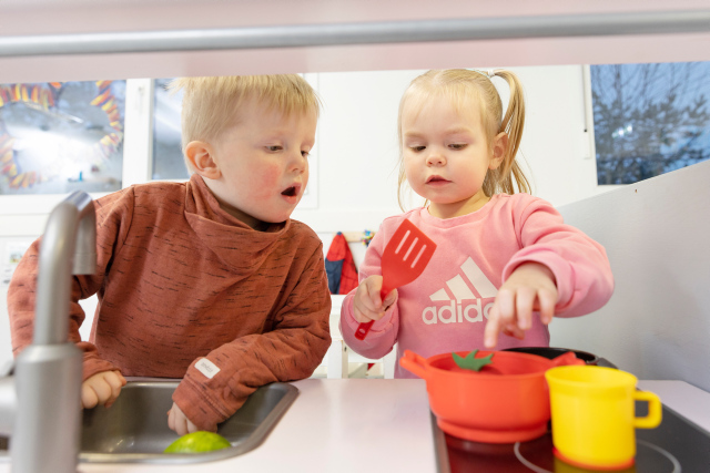 To barn på ca. to år som leker med kasseroller på et lekekjøkken i barnehagen. En gutt til venstre og jente til høyre. 