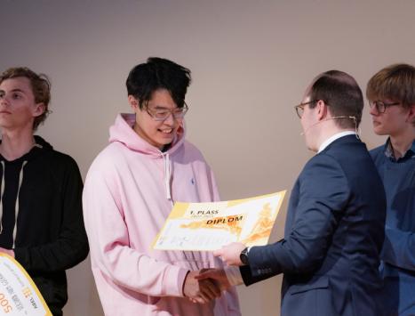 James Jia (18) får utdelt en premiesjekk fra statssekretær i kunnskapsdepartementet, Sindre Lysø. I bakgrunnen ser vi to gutter på ca. 18 år