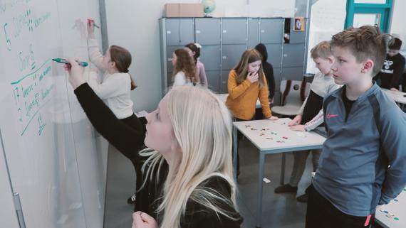 Jente på ca. 10 år skriver på en vertikal tavle. Bak henne står det en gutt og ser på hva hun skriver, og i bakgrunnen ser vi flere andre elever som står og ser på vertikale tavler.