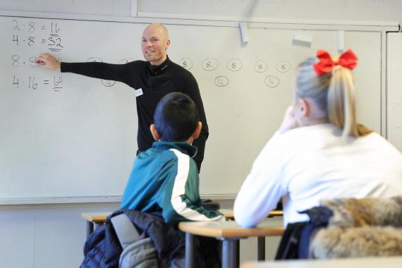 Mann som står ved tavle og underviser. I forgrunnen ser vi elever som sitter med ryggen til.