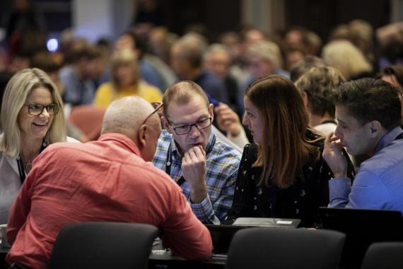 Fem voksne som snakker sammen. Bak ser vi en konferansesal med flere mennesker. 