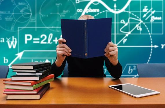 En person som sitter ved et skrivebord og holder en svart bok foran ansiktet. I bakgrunnen ser vi en grønn tavle med matematiske formler.