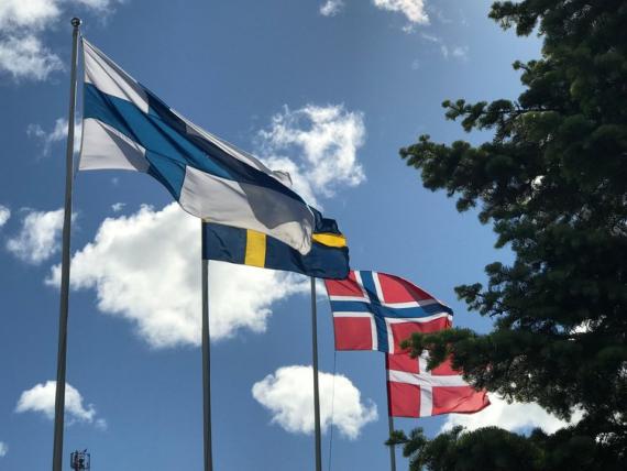 Foto som viser de fire nordiske flaggene heist på flaggstang. I bakgrunnen ser vi blå himmel med skyer.