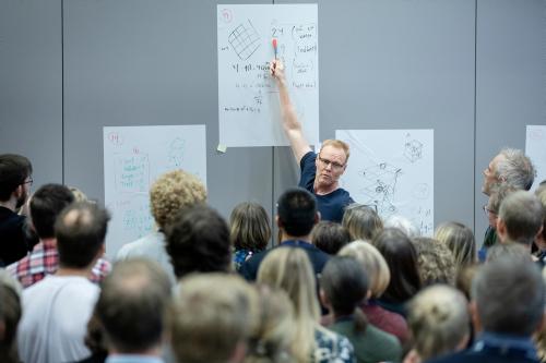 Mann som står foran en gruppe mennesker og peker på en av tre whiteboard-tavler som henger bak ham. Mannen heter Peter Liljedahl. 