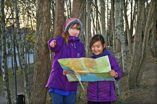 To jenter som står i skogen og ser på et kart. Den ene jenta peker foran seg. 