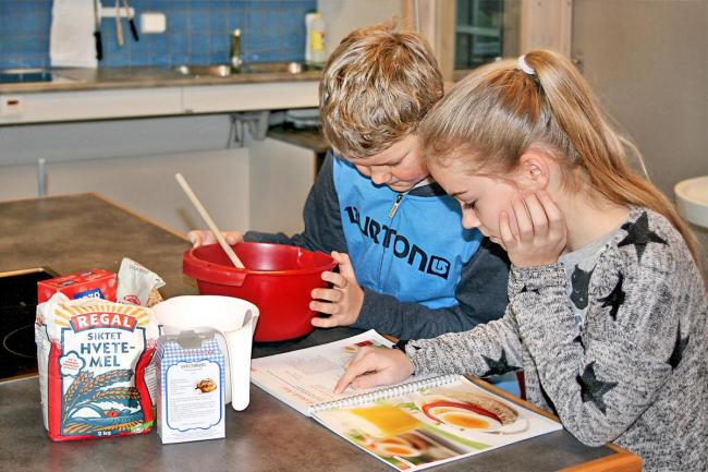 Jente og gutt som sitter på et kjøkkenbord med en kokebok foran seg