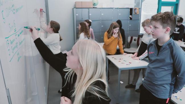 Bilde av flere elever som står og peker på en tavle, i et klasserom. 