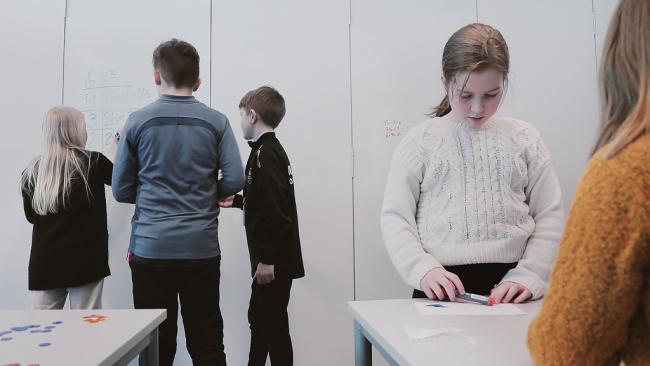 Elever som står i et klasserom. Noen jobber på en tavle ved veggen.