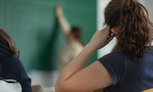 Bilde av jente som sitter i klasserom, med ryggen til. Læreren vises i bakgrunnen