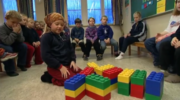 Jente som sitter på golvet i et klasserom med kjempestore legoklosser foran seg. Bak sitter resten av klassen hennes. 