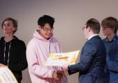 James Jia (18) får utdelt en premiesjekk fra statssekretær i kunnskapsdepartementet, Sindre Lysø. I bakgrunnen ser vi to gutter på ca. 18 år