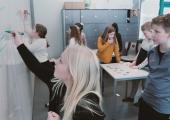Jente på ca. 10 år skriver på en vertikal tavle. Bak henne står det en gutt og ser på hva hun skriver, og i bakgrunnen ser vi flere andre elever som står og ser på vertikale tavler.