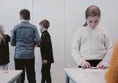 Elever på 5. trinn som står og jobber i et klasserom. Tre elever står rundt et bord, litt lengre borte ser vi tre elever som står ved en vertikal tavle på veggen. 
