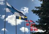 Foto som viser de fire nordiske flaggene heist på flaggstang. I bakgrunnen ser vi blå himmel med skyer.