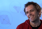 Bilde av en mann i rød genser, sitter foran en blå bakgrunn med matematiske figurer. 
